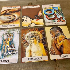 May include: Six tarot cards laid out on a wooden surface. The cards are from left to right: "Spiritual", "Indigenous", "Father", "Female", "Betrayal", and "Good Karma".