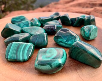 May include: A collection of smooth, polished malachite tumbled stones in various shades of green. The stones have a natural, striated pattern and are arranged on a wooden surface.