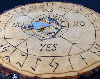 Könnte beinhalten: Ein Holz-Pendelbrett mit Runen und den Worten "YES" und "NO" beschriftet. Ein weißes Opal-Pendel hängt an einer silbernen Kette.