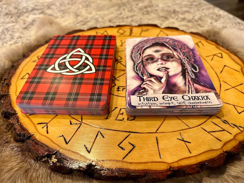 May include: Two tarot cards on a wooden circular base with runes carved into the surface. The card on the left has a red and black plaid pattern with a white Celtic knot design. The card on the right has a colorful illustration of a woman with a third eye and the text "Third Eye Chakra intuition, insight, self-awareness".