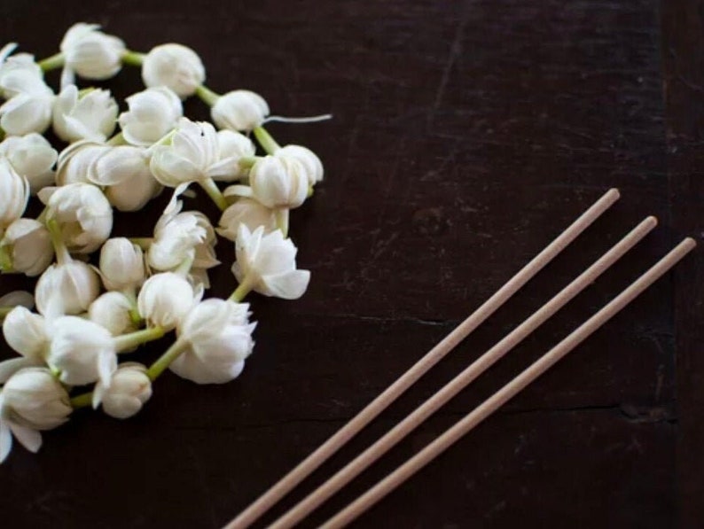 May include: A bunch of white jasmine flowers with green stems on a dark brown wooden surface. Three wooden incense sticks are lying on the surface.