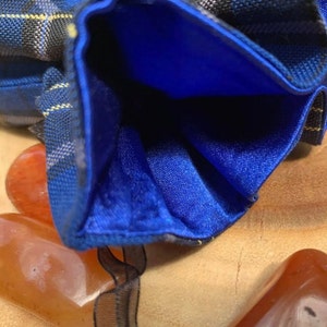 May include: A blue and black plaid fabric pouch with a blue satin lining. The pouch is open and the lining is visible. The pouch is sitting on a wooden surface.