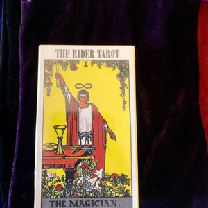 May include: The Rider Tarot card, The Magician, features a yellow background with a man in a red robe holding a wand. The card is surrounded by a border of roses and vines.