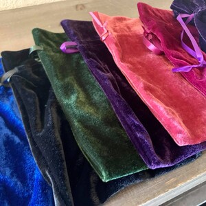 May include: A set of six velvet drawstring pouches in various colors, including blue, black, green, purple, pink, and red. The pouches are arranged in a stack, with the blue pouch at the bottom and the red pouch at the top.