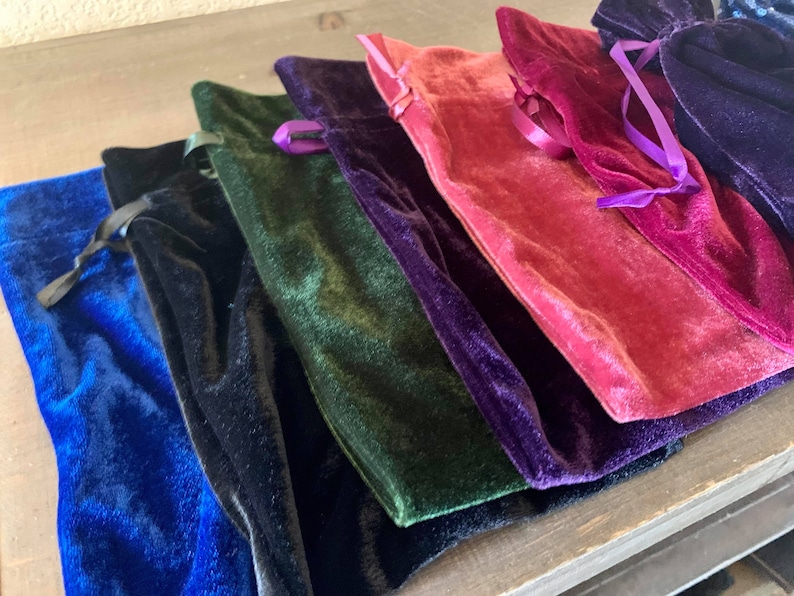 May include: A set of six velvet drawstring pouches in various colors, including blue, black, green, purple, pink, and red. The pouches are arranged in a stack, with the blue pouch at the bottom and the red pouch at the top.