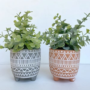 May include: Two potted plants with green leaves in geometric patterned pots. The pot on the left is gray with white geometric designs. The pot on the right is orange with white geometric designs.