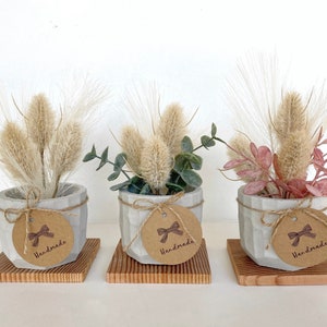 May include: Three small concrete planters with a geometric design, each filled with dried grasses and greenery. The planters have a rustic brown paper tag tied around them with twine, each tag has a brown ribbon and the word "Handmade" printed on it.
