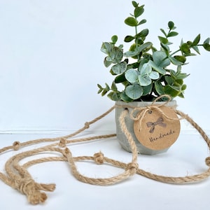 May include: A small concrete planter with a hanging rope, filled with a green leafy plant. The planter has a brown paper tag with a bow and the word "Handmade" written on it.