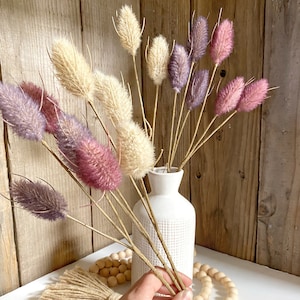 Puede incluir: Un jarrón de cerámica blanca con superficie texturizada contiene un ramo de flores secas. Las flores son una mezcla de tonos beige, morado y rosa. El jarrón está sobre una superficie blanca con una guirnalda de cuentas de madera.