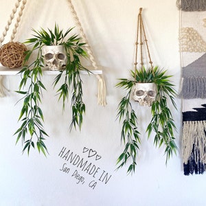 Puede incluir: Dos macetas de calavera blancas con plantas verdes frondosas colgando de una cuerda de macramé. El texto "HANDMADE IN San Diego, CA" está escrito en una pared blanca.