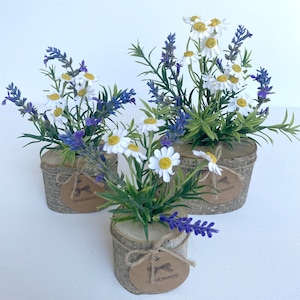 May include: Three small wooden pots with a rustic bark texture, each containing a bouquet of white daisies and purple lavender. The pots are tied with twine and have a brown paper tag that reads "HANDMADE".