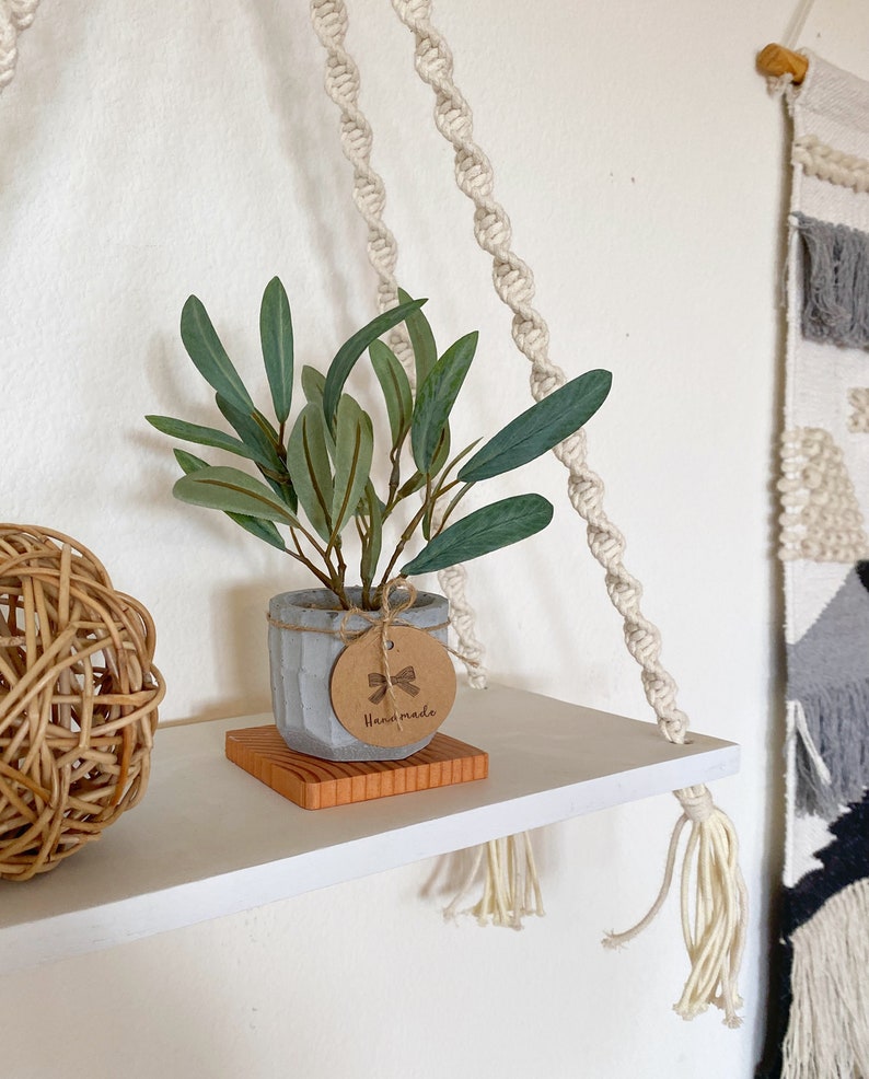 May include: A white shelf with a small potted plant, a woven ball, and a macrame hanging. The plant is in a grey concrete pot with a brown tag that says "Handmade".