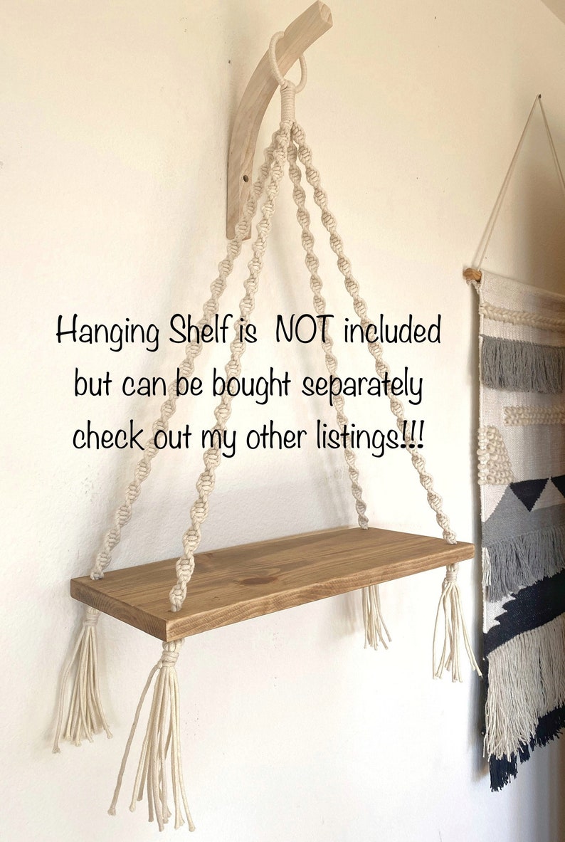 Puede incluir: Una estanter&iacute;a colgante de macram&eacute; con una repisa de madera y borlas blancas. La estanter&iacute;a est&aacute; colgada de un soporte de madera fijado a una pared. El texto "Hanging Shelf is NOT included but can be bought separately check out my other listings!!!" es visible en la imagen.