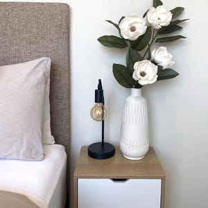 May include: A bedroom nightstand with a black lamp and a white vase holding white flowers and green leaves. The nightstand has a light wood top and two white drawers. A bed with a beige headboard and pillow is in the background.