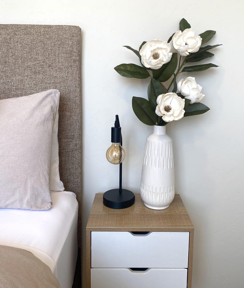 May include: A bedroom nightstand with a black lamp and a white vase holding white flowers and green leaves. The nightstand has a light wood top and two white drawers. A bed with a beige headboard and pillow is in the background.