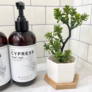 May include: A brown glass bottle with a black pump dispenser filled with hand wash labeled "No. 7 Cypress hand wash 8 fl oz / 237 ml Magnolia Home 2002". A white hexagonal ceramic planter with a small green succulent plant sits on a wooden hexagonal coaster.