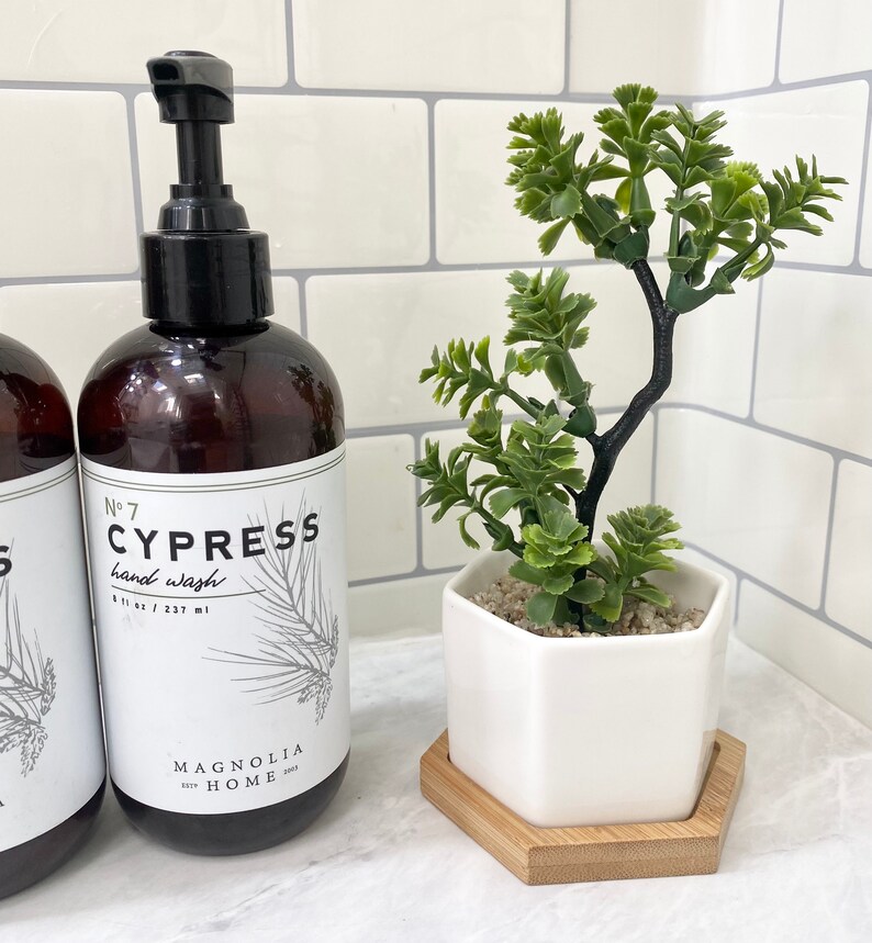 May include: A brown glass bottle with a black pump dispenser filled with hand wash labeled "No. 7 Cypress hand wash 8 fl oz / 237 ml Magnolia Home 2002". A white hexagonal ceramic planter with a small green succulent plant sits on a wooden hexagonal coaster.