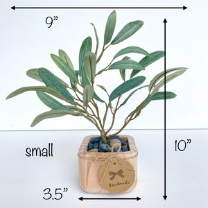 May include: A small artificial olive tree in a wooden pot with a brown tag that says "Handmade". The tree is 9 inches wide and 10 inches tall. The pot is 3.5 inches wide.