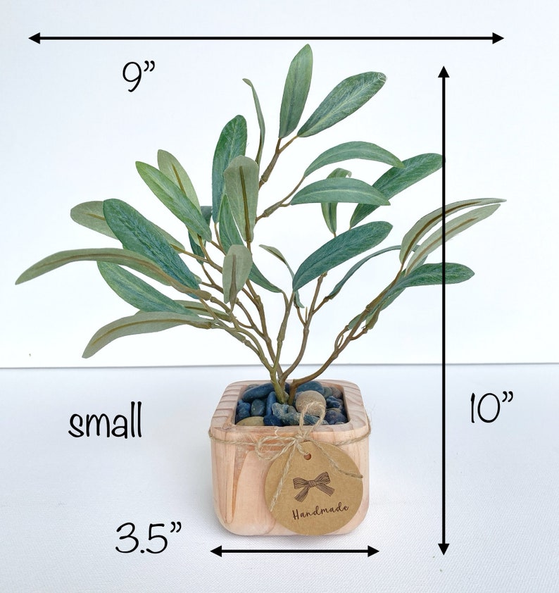 May include: A small artificial olive tree in a wooden pot with a brown tag that says "Handmade". The tree is 9 inches wide and 10 inches tall. The pot is 3.5 inches wide.