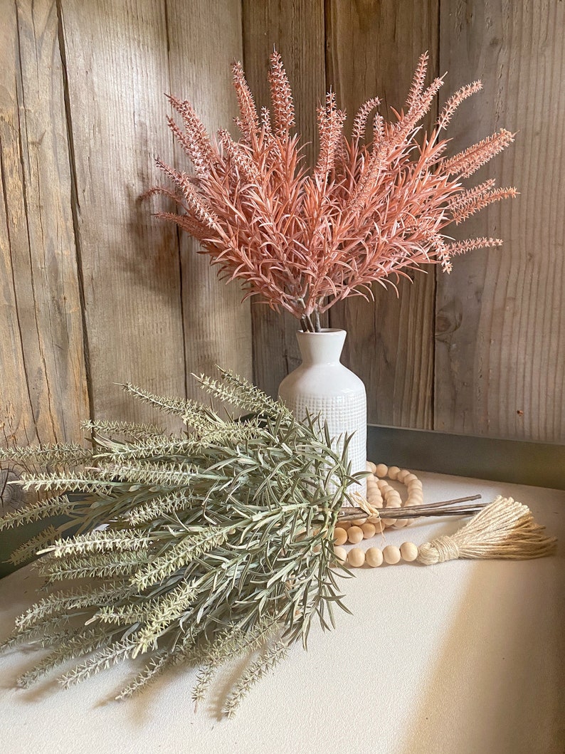 May include: Two bouquets of faux lavender in a white vase and on a table with a wooden bead garland. The lavender on the left is a light green color and the lavender on the right is a dusty rose color.