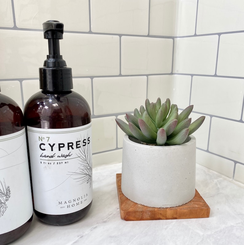 May include: A brown hand wash bottle with a black pump and a white label that reads "No 7 CYPRESS hand wash" with a pine branch illustration. A small succulent plant in a white concrete pot sits on a wooden square.