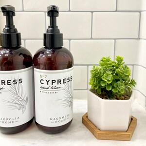 May include: Two amber glass bottles with black pump tops and white labels. The labels read "No. 7 Cypress hand lotion 8 fl oz / 237 ml" and "Cypress wash". Both bottles have a pine tree design and the text "Magnolia Home" at the bottom. A small white hexagonal planter with a green succulent plant is in the foreground.