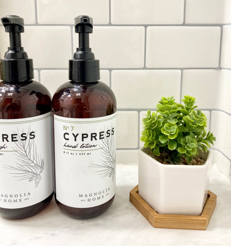 May include: Two amber glass bottles with black pump tops and white labels. The labels read "No. 7 Cypress hand lotion 8 fl oz / 237 ml" and "Cypress wash". Both bottles have a pine tree design and the text "Magnolia Home" at the bottom. A small white hexagonal planter with a green succulent plant is in the foreground.