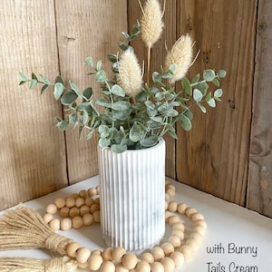 May include: A white marble vase with a ribbed texture holds a bouquet of dried beige flowers and green eucalyptus leaves. A strand of wooden beads with tassels sits in front of the vase on a white surface. The text "with Bunny Tails Cream" is visible in the bottom right corner of the image.