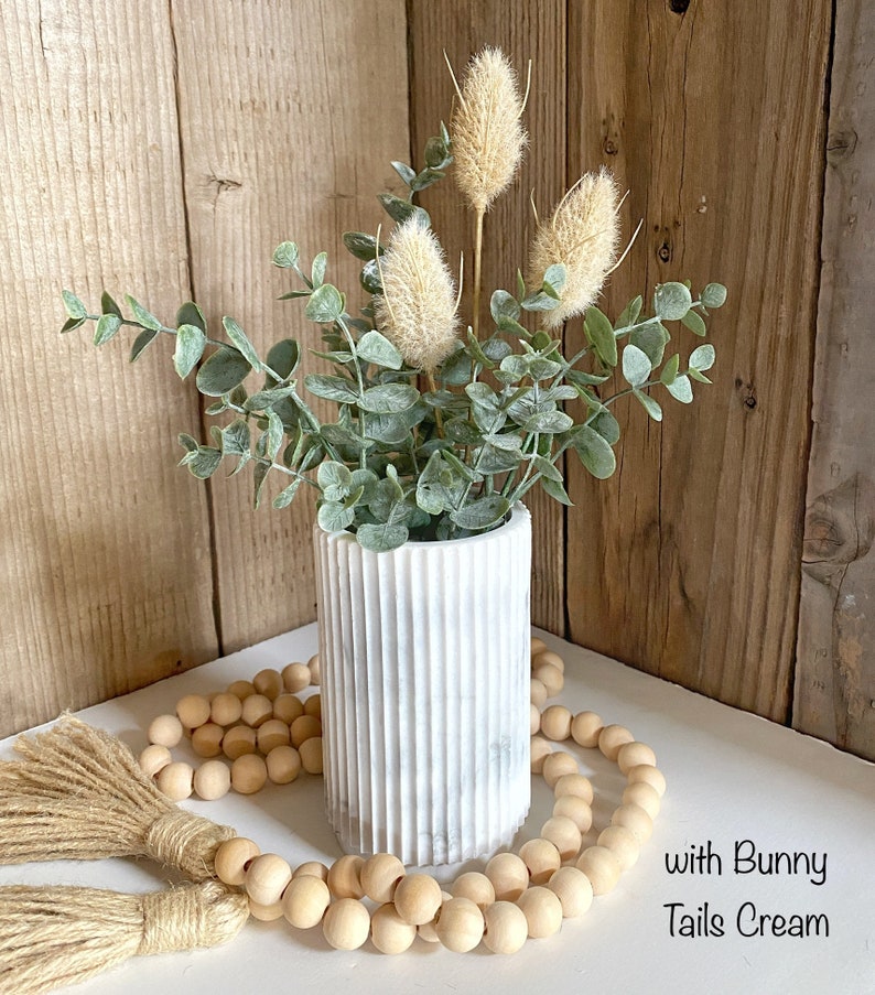 May include: A white marble vase with a ribbed texture holds a bouquet of dried beige flowers and green eucalyptus leaves. A strand of wooden beads with tassels sits in front of the vase on a white surface. The text "with Bunny Tails Cream" is visible in the bottom right corner of the image.