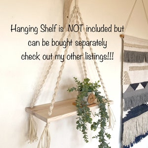 May include: A macrame plant hanger with a wooden shelf and tassels. The shelf is empty, but a potted plant is hanging from the hanger. The text "Hanging Shelf is NOT included but can be bought separately check out my other listings!!!" is visible in the image.