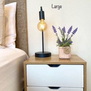 K&ouml;nnte beinhalten: Ein Nachttisch mit zwei Schubladen, einer schwarzen Lampe mit einer Gl&uuml;hbirne und einer Topfpflanze mit lila Bl&uuml;ten. Der Tisch ist aus Holz mit einer hellbraunen Oberfl&auml;che. Die Lampe hat einen schwarzen Metallfu&szlig; und einen schwarzen Metallschirm. Die Pflanze steht in einem rosafarbenen Keramiktopf mit einem braunen Papieretikett, auf dem "Handmade" steht. Der Text "Large" ist an der Wand hinter dem Tisch geschrieben.