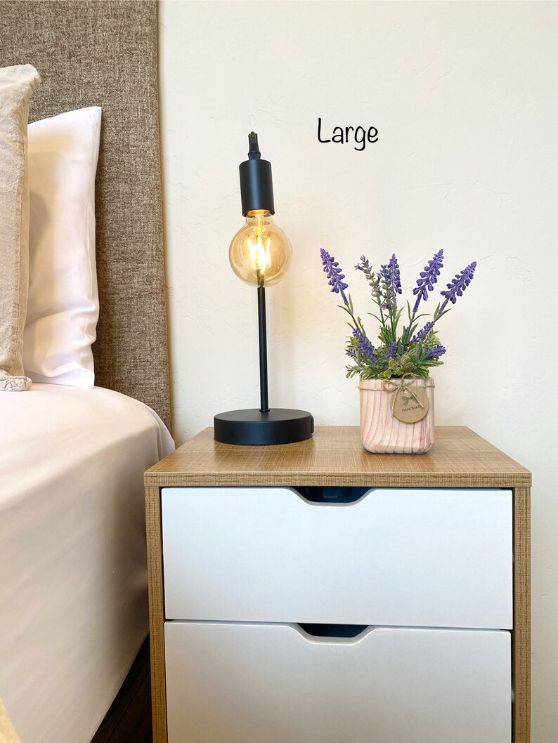 K&ouml;nnte beinhalten: Ein Nachttisch mit zwei Schubladen, einer schwarzen Lampe mit einer Gl&uuml;hbirne und einer Topfpflanze mit lila Bl&uuml;ten. Der Tisch ist aus Holz mit einer hellbraunen Oberfl&auml;che. Die Lampe hat einen schwarzen Metallfu&szlig; und einen schwarzen Metallschirm. Die Pflanze steht in einem rosafarbenen Keramiktopf mit einem braunen Papieretikett, auf dem "Handmade" steht. Der Text "Large" ist an der Wand hinter dem Tisch geschrieben.
