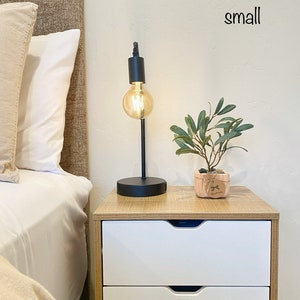 May include: A small wooden nightstand with two white drawers. A black table lamp with a round, clear glass bulb is on the top of the nightstand. A small potted plant with green leaves is on the nightstand next to the lamp. The word "small" is written on the wall above the nightstand.