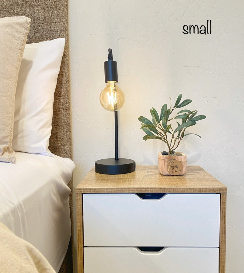 May include: A small wooden nightstand with two white drawers. A black table lamp with a round, clear glass bulb is on the top of the nightstand. A small potted plant with green leaves is on the nightstand next to the lamp. The word "small" is written on the wall above the nightstand.