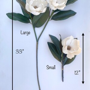 May include: Two artificial magnolia flower stems. The large stem is 33 inches long and features three white flowers and green leaves. The small stem is 12 inches long and has one flower and leaves. The flowers have a brown center.