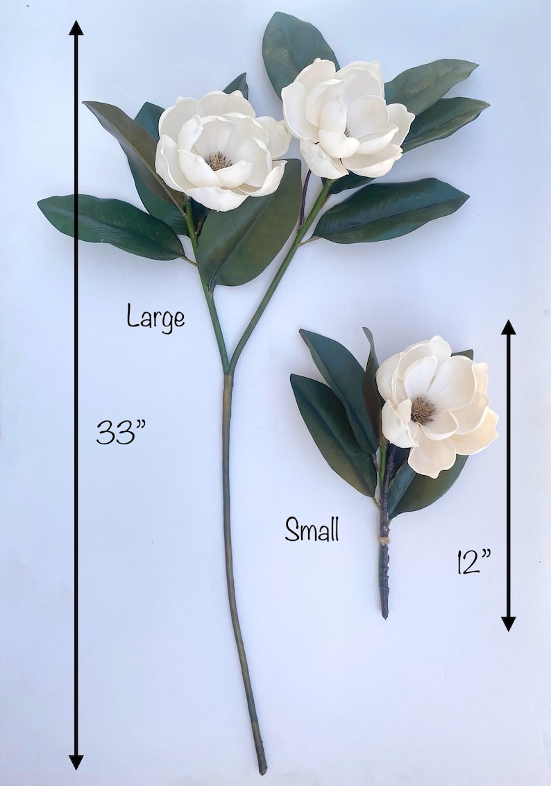 May include: Two artificial magnolia flower stems. The large stem is 33 inches long and features three white flowers and green leaves. The small stem is 12 inches long and has one flower and leaves. The flowers have a brown center.