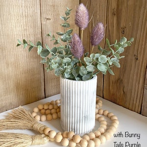 May include: A white marble vase with a ribbed texture holds a bouquet of dried eucalyptus and purple bunny tail flowers. A strand of wooden beads with tassels is in the foreground. The text "with Bunny Tails Purple" is visible in the lower right corner of the image.