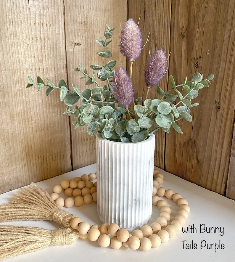 May include: A white marble vase with a ribbed texture holds a bouquet of dried eucalyptus and purple bunny tail flowers. A strand of wooden beads with tassels is in the foreground. The text "with Bunny Tails Purple" is visible in the lower right corner of the image.