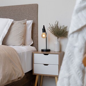 May include: A bedside table with two drawers and a lamp with a light bulb turned on. The table has wooden legs and is next to a bed with a brown headboard and white bedding.