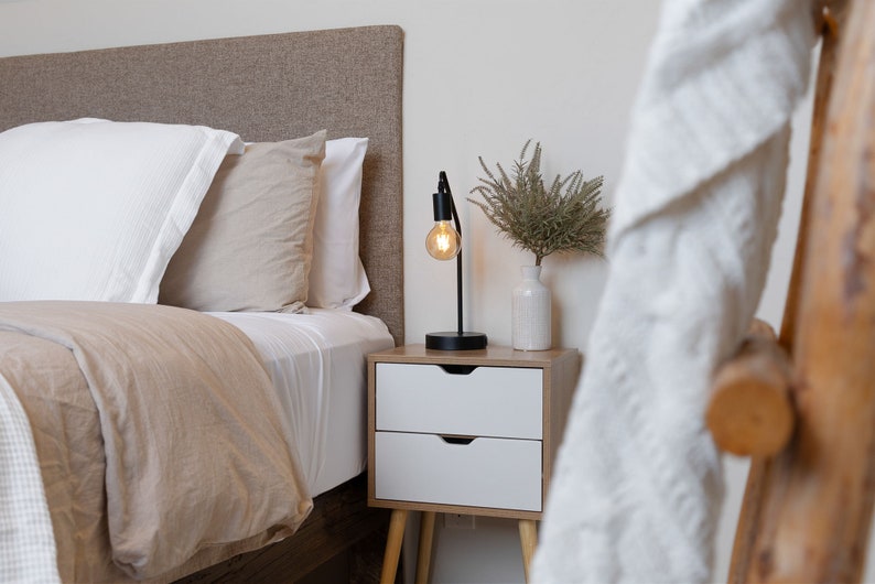 May include: A bedside table with two drawers and a lamp with a light bulb turned on. The table has wooden legs and is next to a bed with a brown headboard and white bedding.