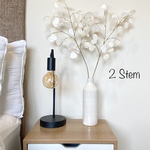 May include: Two white stems of dried flowers in a white textured vase on a wooden nightstand with a black lamp with a light bulb. The text "2 Stem" is written on the wall.