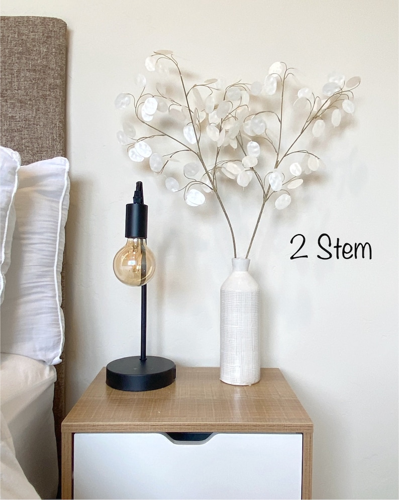 May include: Two white stems of dried flowers in a white textured vase on a wooden nightstand with a black lamp with a light bulb. The text "2 Stem" is written on the wall.