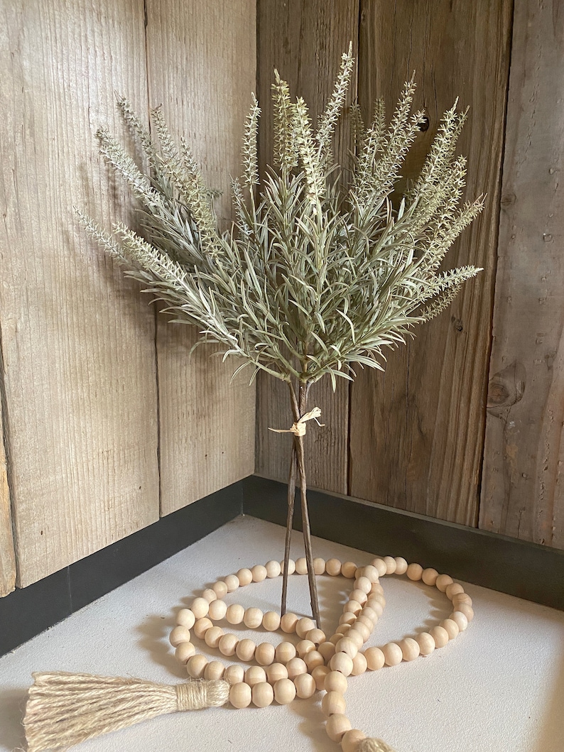 May include: A bouquet of dried, light green, artificial lavender stems with a wooden bead garland with a jute tassel.