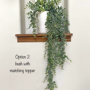 May include: A white ceramic vase with a leafy green plant cascading down from it. The vase is sitting on a wooden shelf. The text "Option 2 bush with matching topper" is written below the shelf.