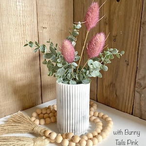 May include: White marble vase with a ribbed texture, filled with a bouquet of pink bunny tail grass and eucalyptus leaves. The vase is sitting on a white surface with a wooden bead garland and a brown tassel.