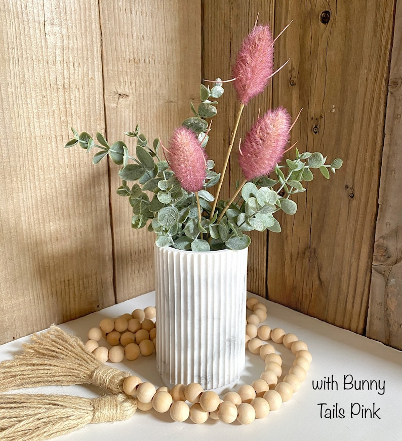 May include: White marble vase with a ribbed texture, filled with a bouquet of pink bunny tail grass and eucalyptus leaves. The vase is sitting on a white surface with a wooden bead garland and a brown tassel.
