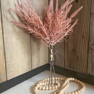 May include: A bouquet of blush pink faux lavender stems with brown wooden stems, arranged in a corner with a wooden bead garland.