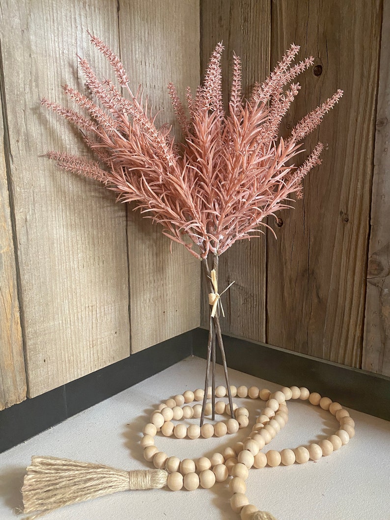 May include: A bouquet of blush pink faux lavender stems with brown wooden stems, arranged in a corner with a wooden bead garland.