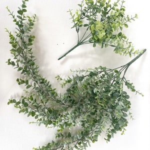 May include: Two bunches of artificial greenery, one with long trailing stems and the other with shorter stems. The greenery is a light green color and has a realistic appearance.