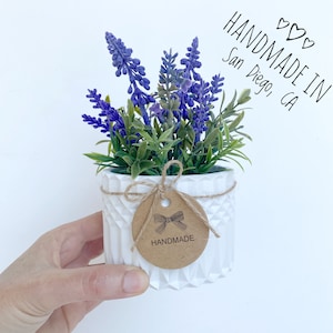 May include: A small white planter with a geometric pattern, filled with purple lavender flowers. A brown paper tag with a handwritten "HANDMADE" label is tied to the planter with twine. The tag also has a small illustration of a bow. The text "HANDMADE IN San Diego, CA" is written in black marker on a white background.